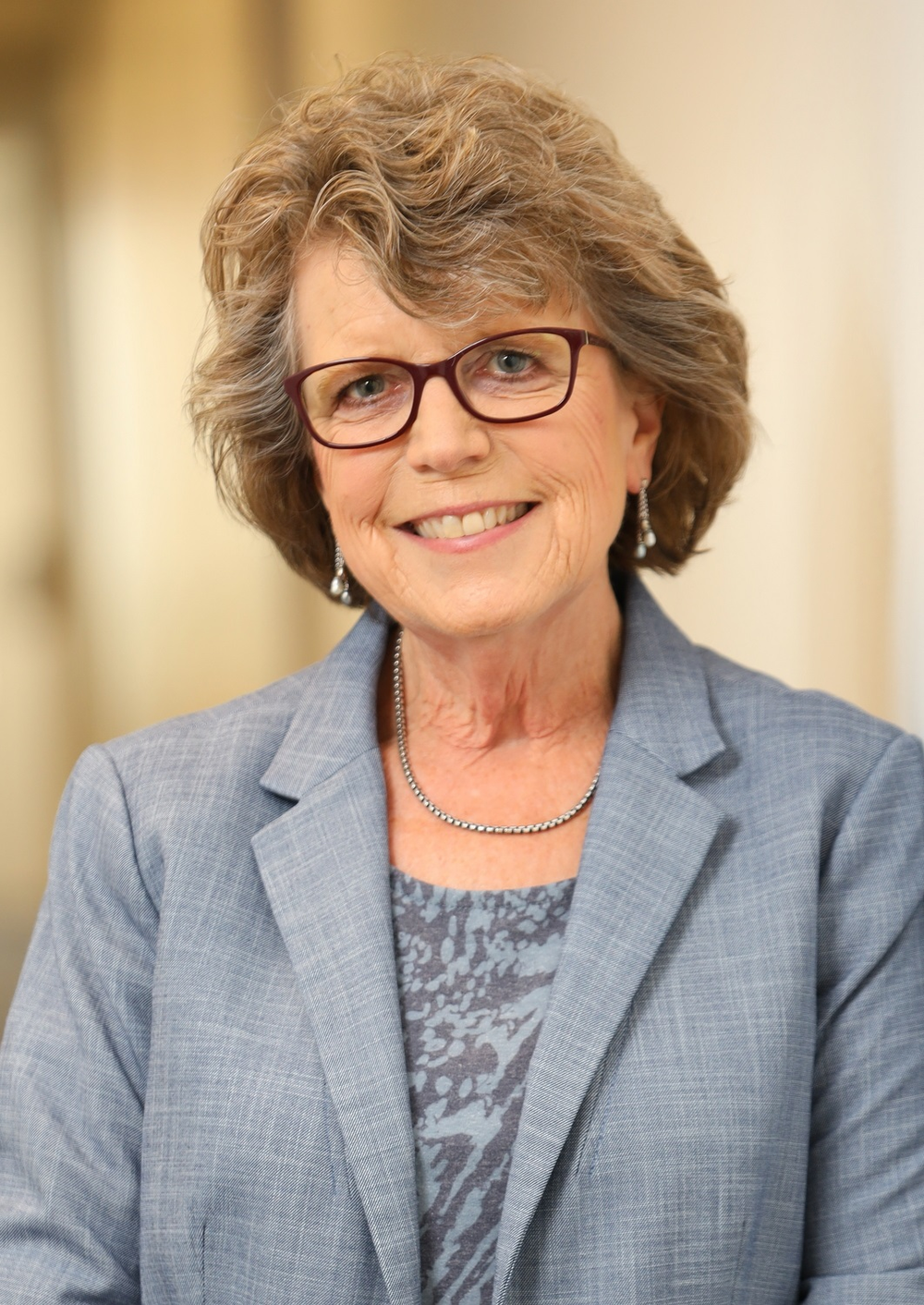 A portrait of a woman with short, curly hair, smiling and wearing glasses and a light gray blazer over a patterned blouse. 